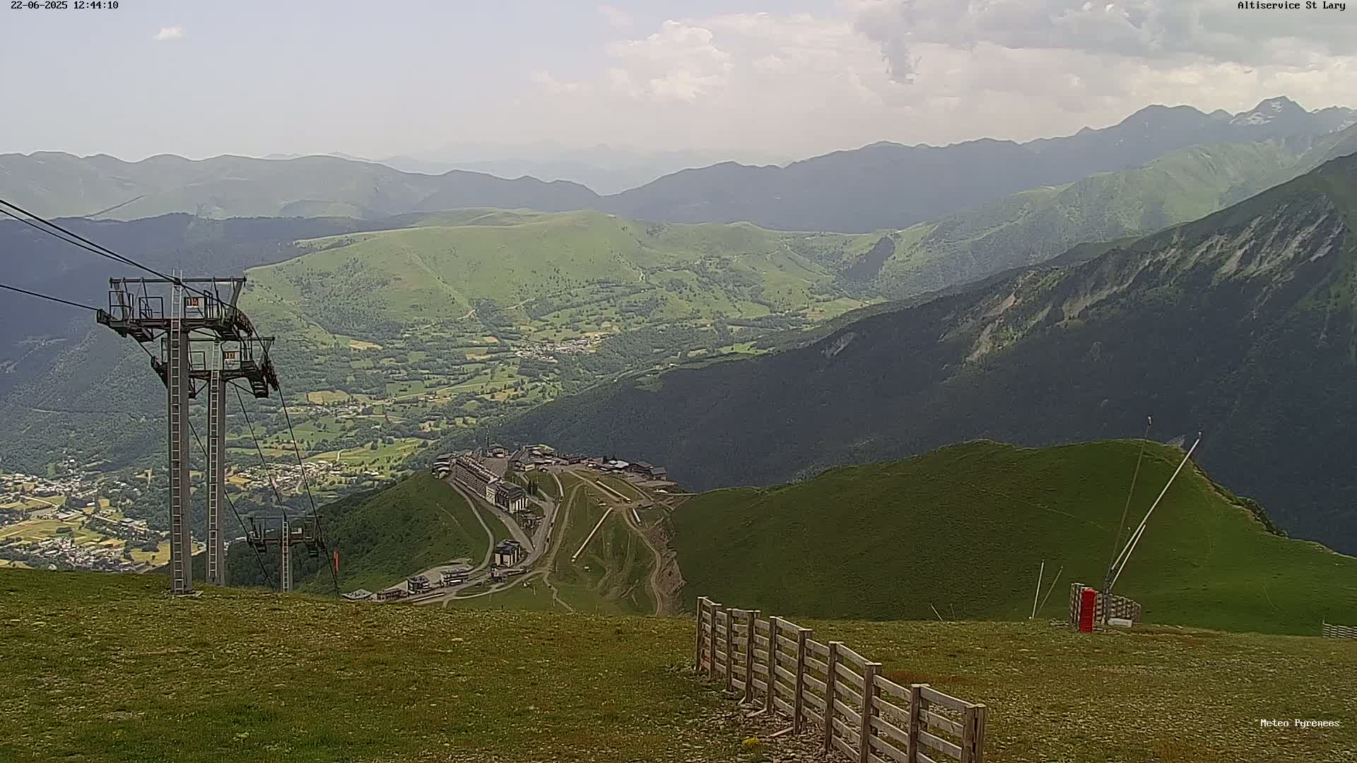 A mountaintop view on a partly cloudy day shows a chairlift, a wooden fence, and a valley with green hills and a village below.