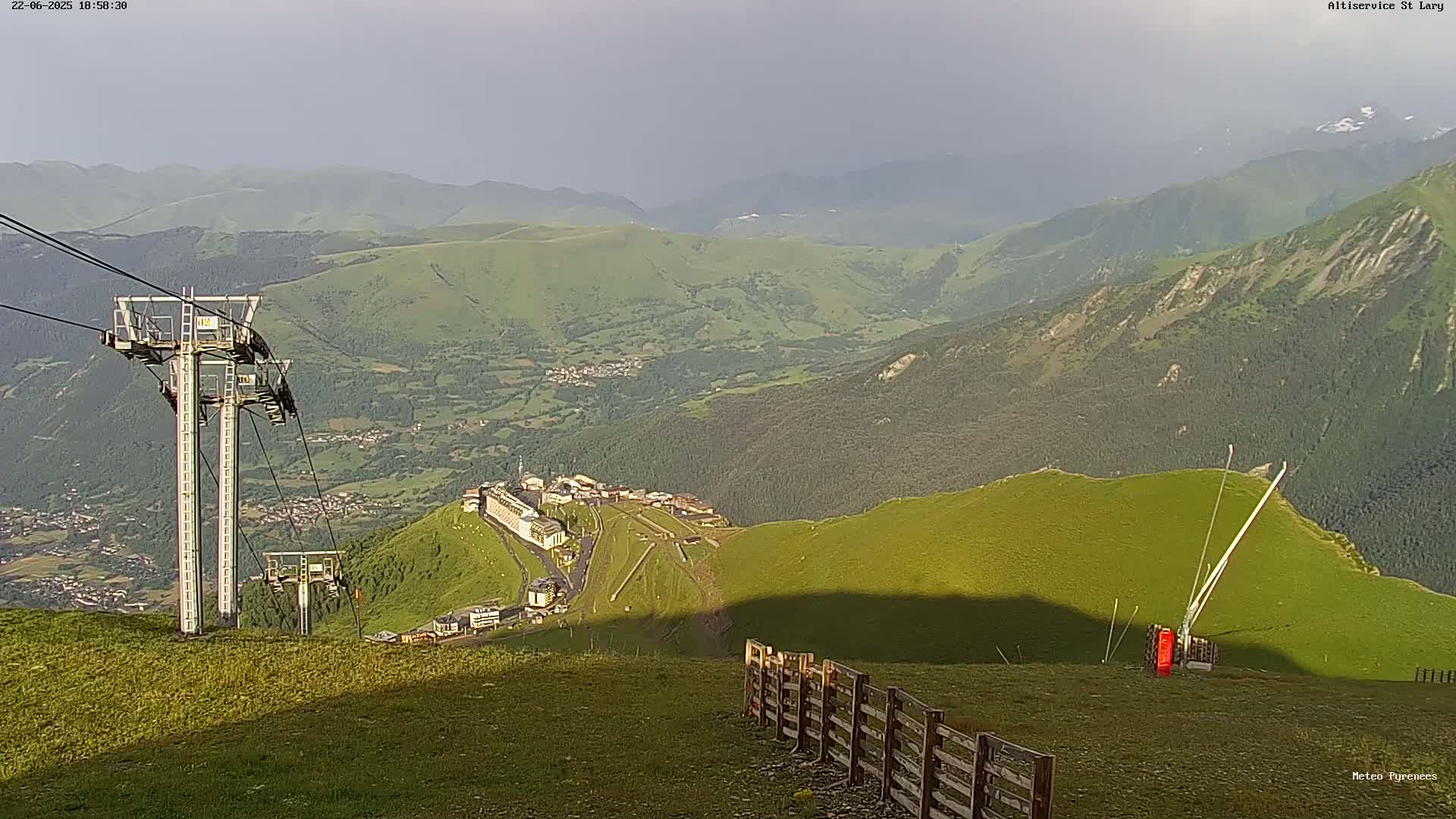 A mountaintop view shows a ski resort nestled in a valley below, with chairlifts visible in the foreground under an overcast sky.