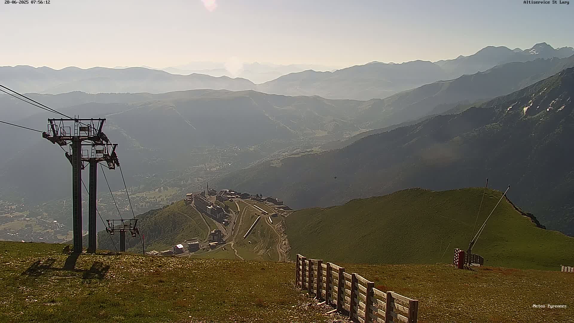A mountaintop view shows a chairlift, a wooden fence, and a hazy valley with distant mountains under a mostly clear sky.