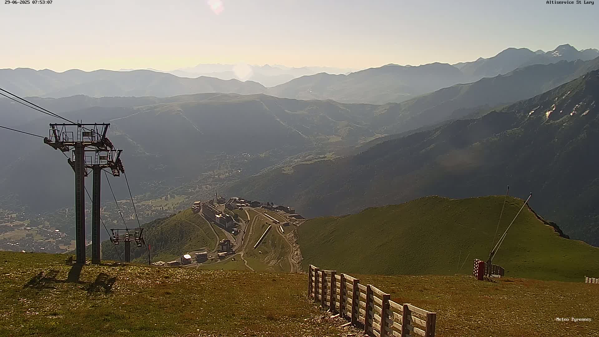 A mountaintop view on a hazy, sunny day shows a chairlift, a wooden fence, and a valley with a village below.