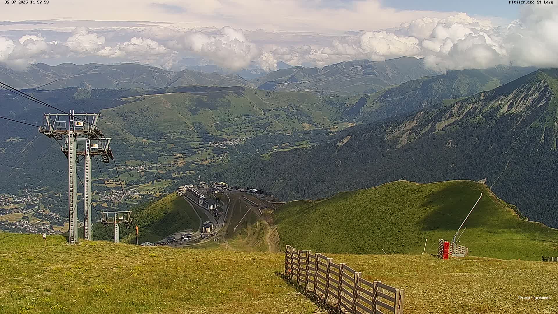 A mountaintop view on a partly cloudy day shows a valley with green hills, a village, and ski resort buildings below, with chairlift towers and a wooden fence in the foreground.
