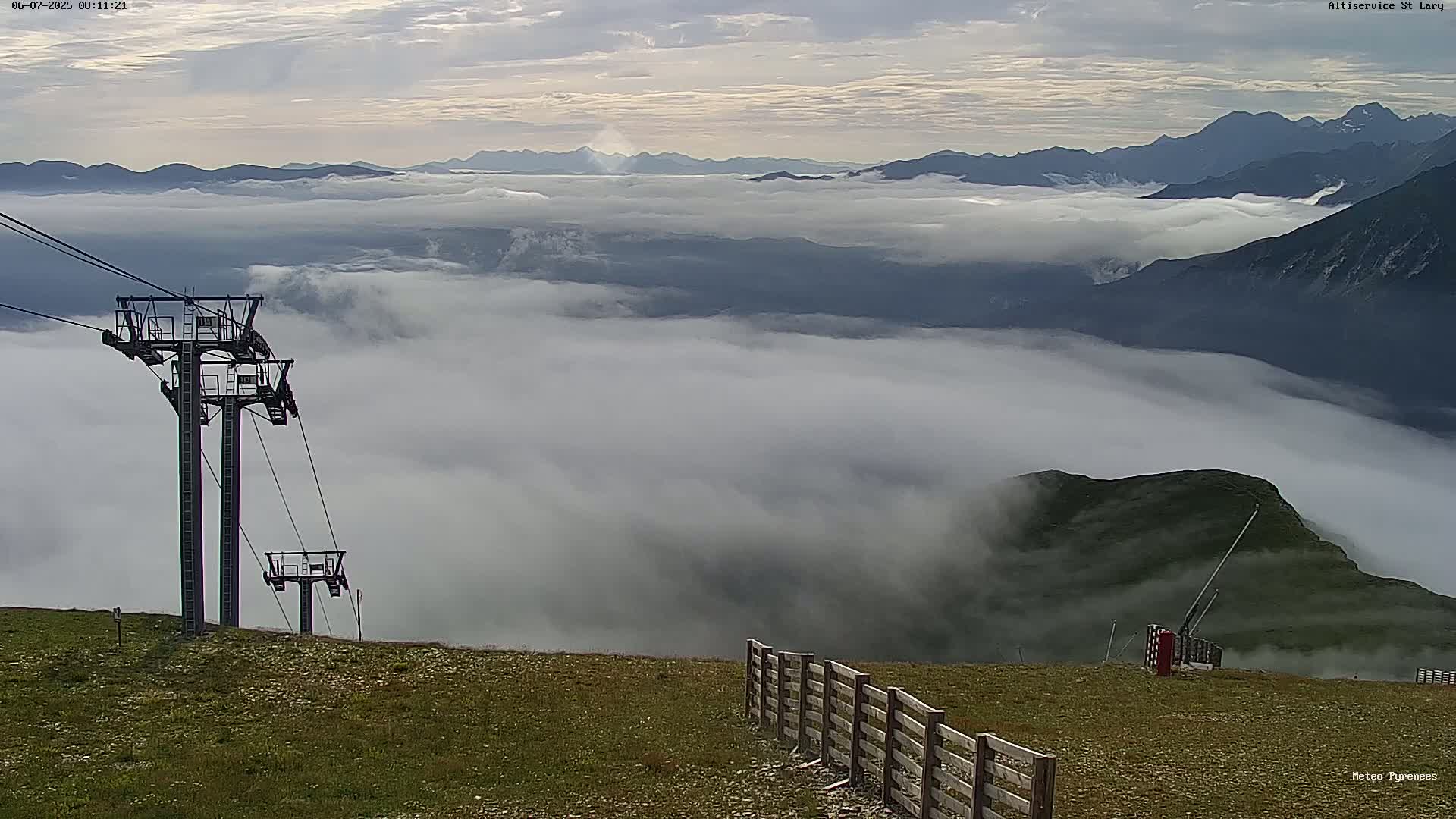 A mountaintop scene shows a chairlift, a wooden fence, and a vast expanse of low-lying clouds below, with distant mountains visible above the clouds under a partly cloudy sky.