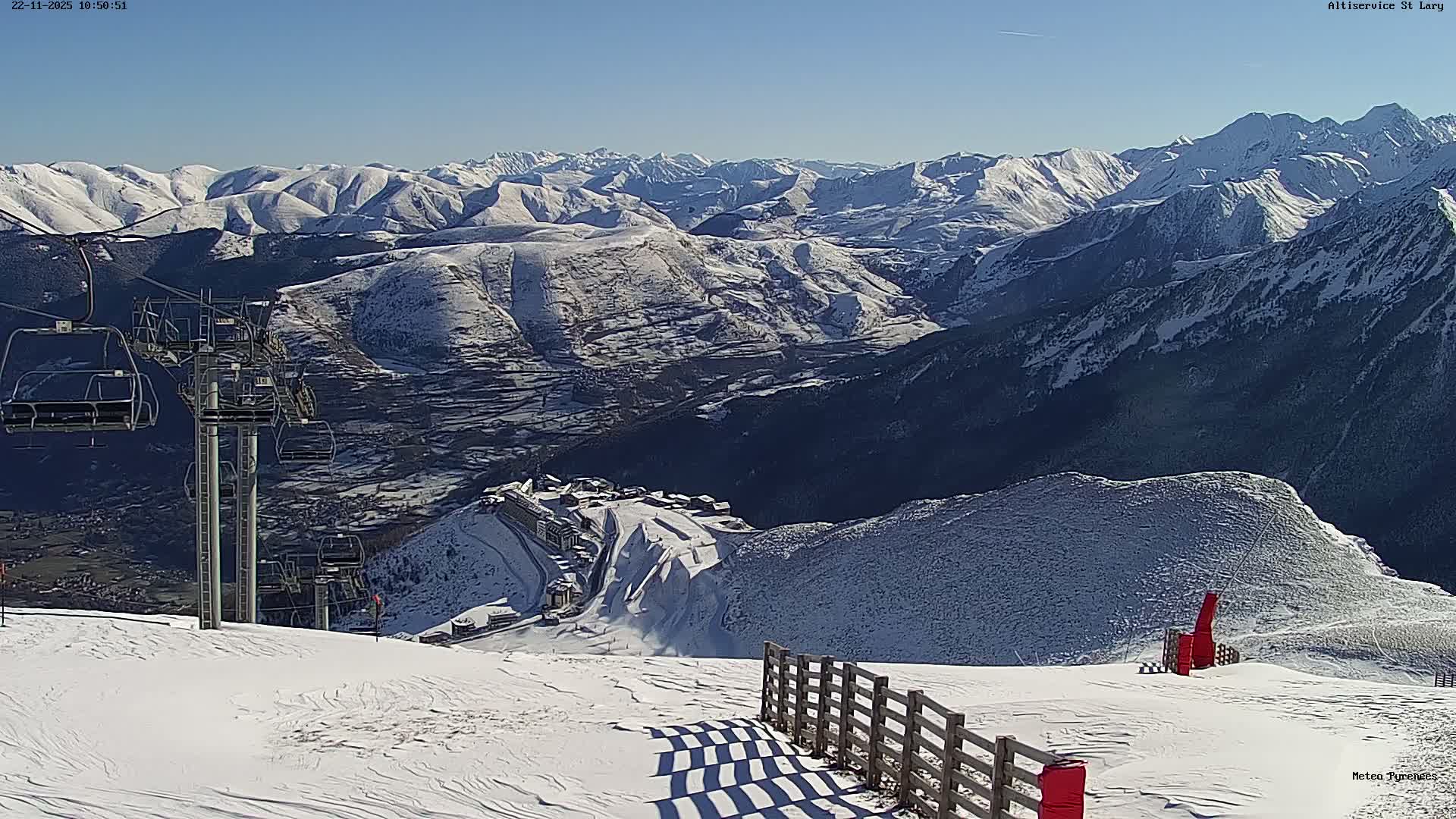 A panoramic view showcases a snow-covered mountain ski resort with chairlifts and buildings, set against a backdrop of distant peaks under a clear, sunny blue sky.