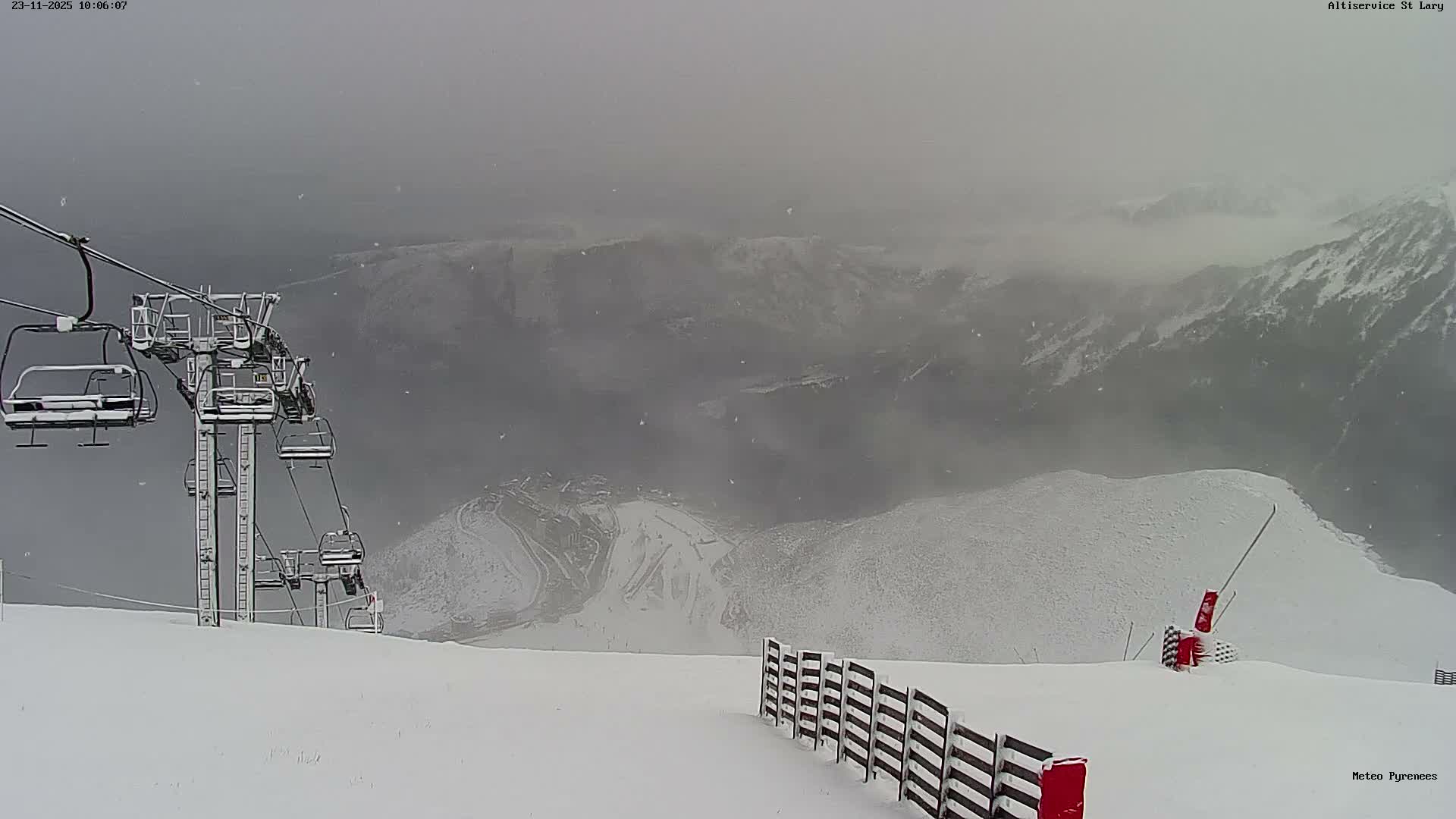 A snowy ski slope with a chairlift on the left and a snow fence on the right descends towards a distant, hazy valley and mountains, all under heavy snowfall and overcast, foggy conditions.