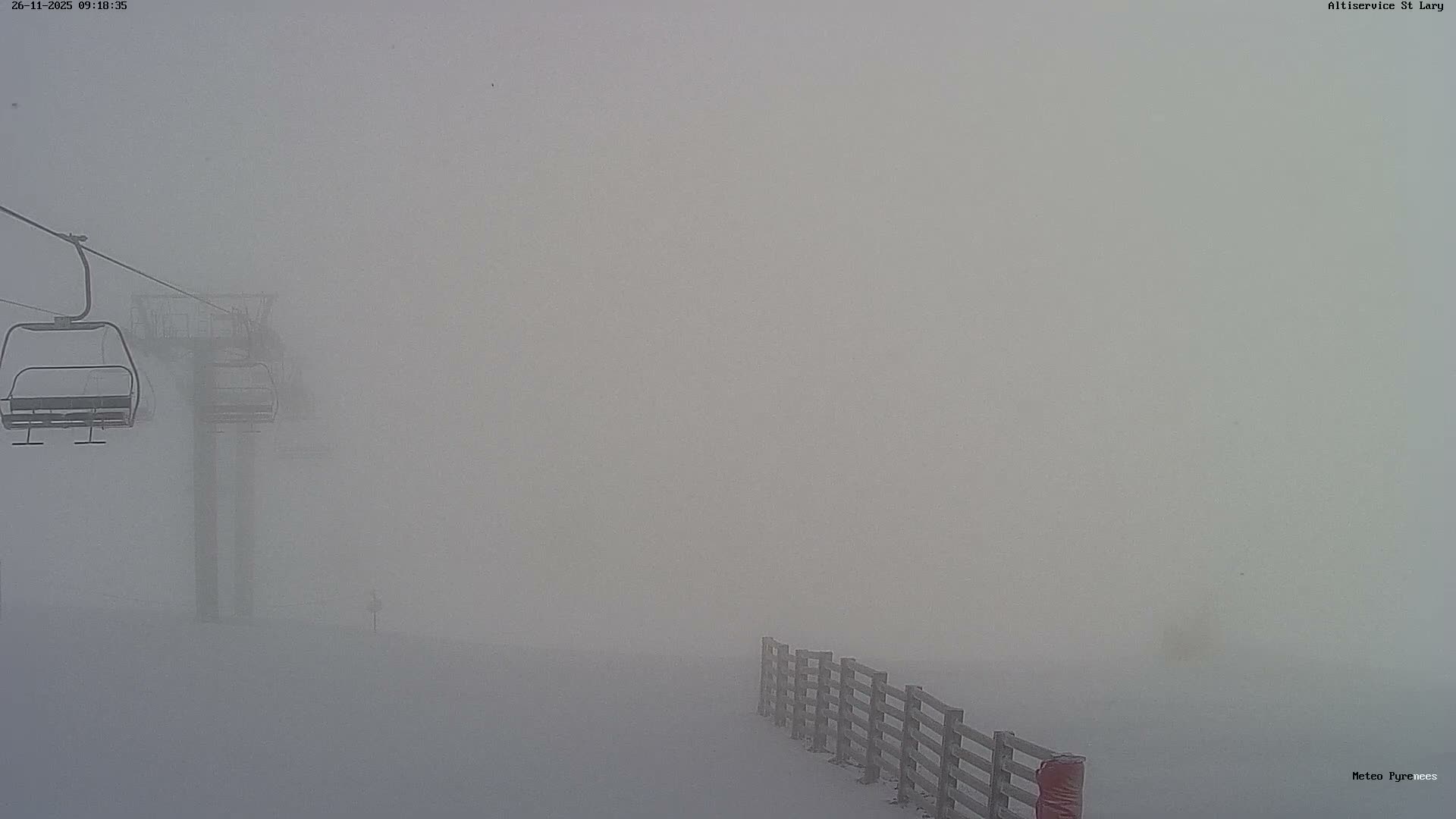 A snow-covered ski slope, featuring empty ski lift chairs and a wooden fence, is enveloped in dense fog or mist, resulting in very low visibility.
