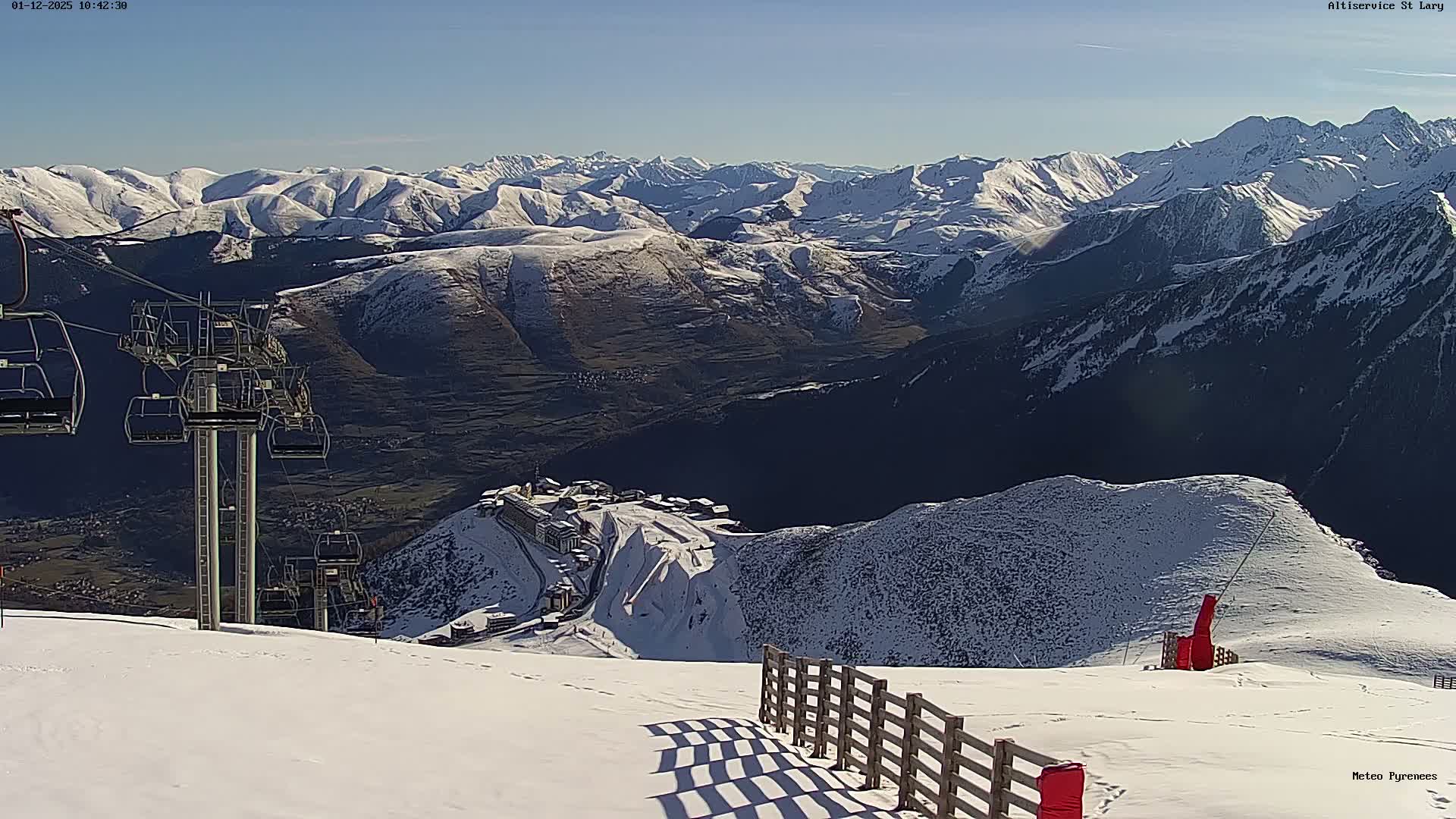 A sunny day reveals a vast snow-covered mountain ski resort landscape, featuring a ski slope with a wooden fence casting long shadows in the foreground, a large ski lift with empty chairs on the left, and a cluster of buildings on a distant snowy peak overlooking a valley, all under a clear blue sky.