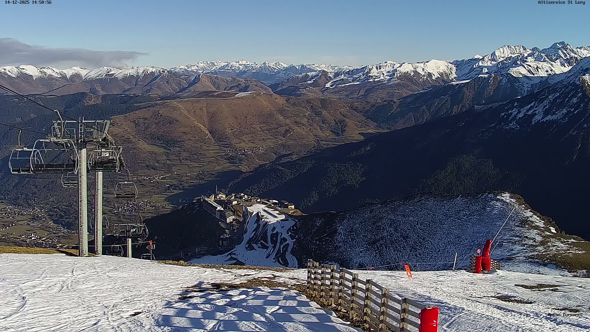 A panoramic view reveals a snow-covered mountain ski resort under an overcast sky with patches of brighter light, featuring chairlifts, distant resort buildings, and a snow cannon actively operating on the slopes.