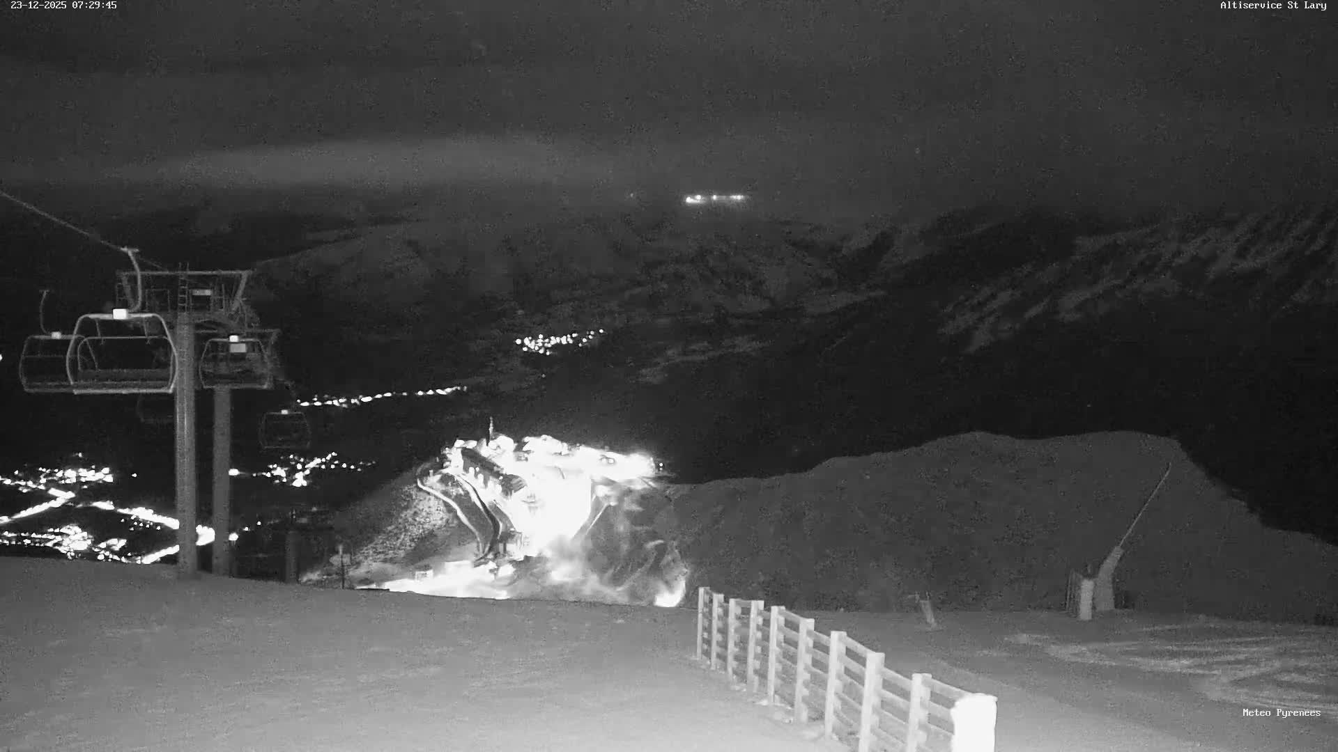 A cold, dark night at a snowy ski resort features a ski lift, distant town lights, and a prominent plume of actively made snow on the slopes.