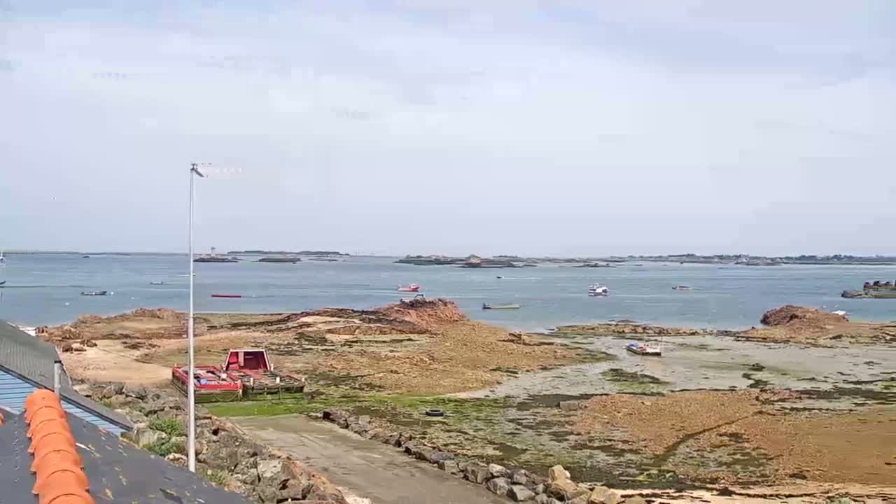 A mostly sunny day reveals a rocky shoreline with several small boats in a calm body of water, distant islands visible in the background.
