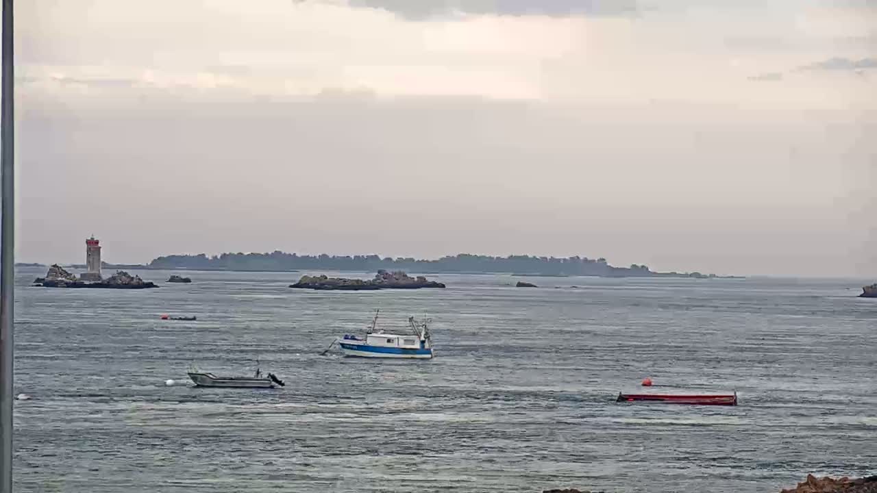 A lighthouse stands on a rocky island in a calm, overcast sea, with several small boats and a larger fishing vessel visible in the distance.