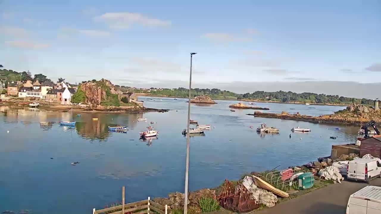 A calm bay with numerous small boats is surrounded by rocky shores and a small village under a mostly clear sky.