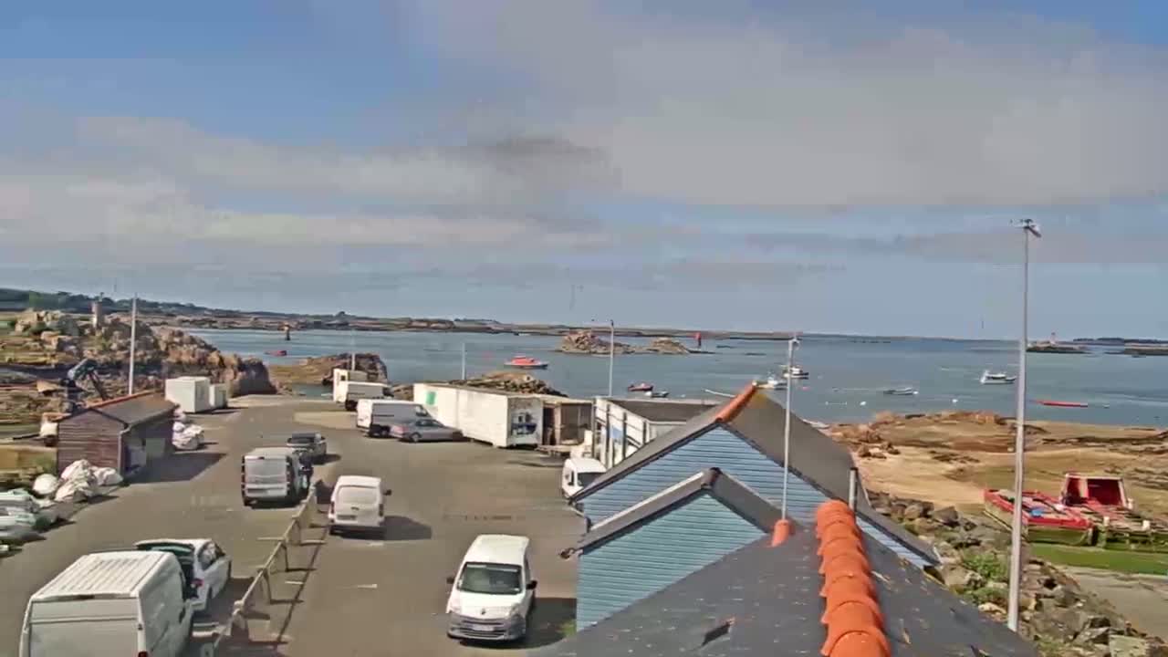 A parking lot with several vans and cars is situated near a waterfront with calm water and small boats under a partly cloudy sky.