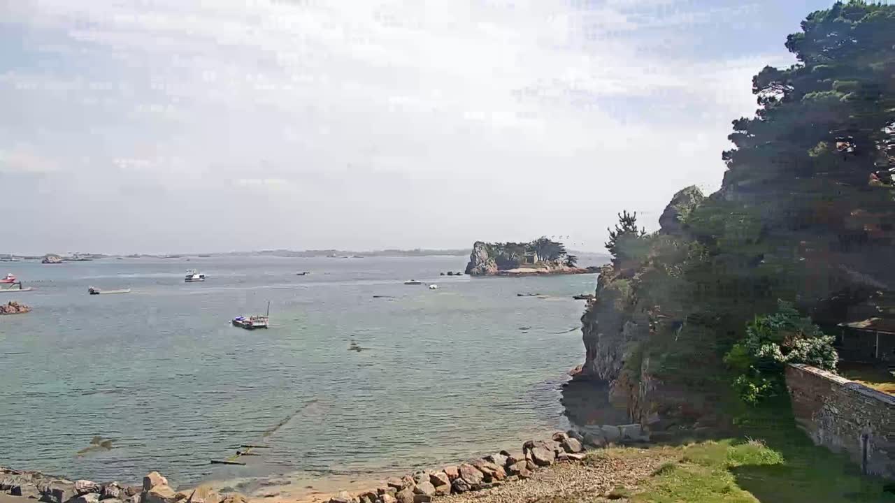 A calm, partly cloudy day shows a rocky shoreline with several small boats in a bay, and a small island in the distance.