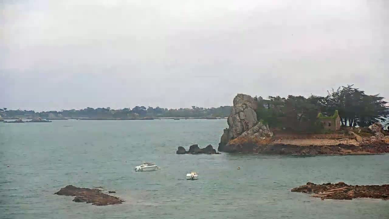 A rocky islet with a small building and trees sits in calm, gray water under an overcast sky, with two small boats nearby and a distant shoreline visible.