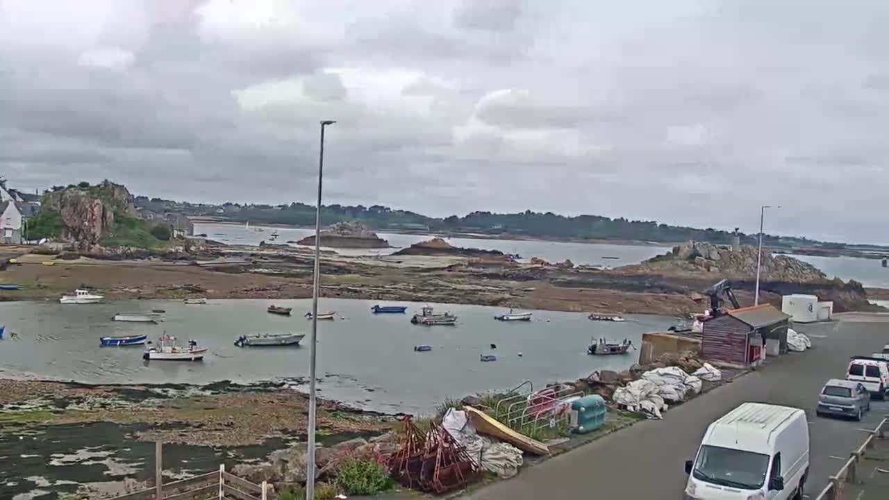 Several small boats are moored in a calm bay under an overcast sky, with a rocky shoreline and distant land visible.