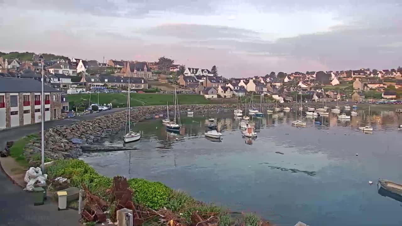 A calm harbor filled with sailboats is nestled beside a hillside town under a cloudy, twilight sky.