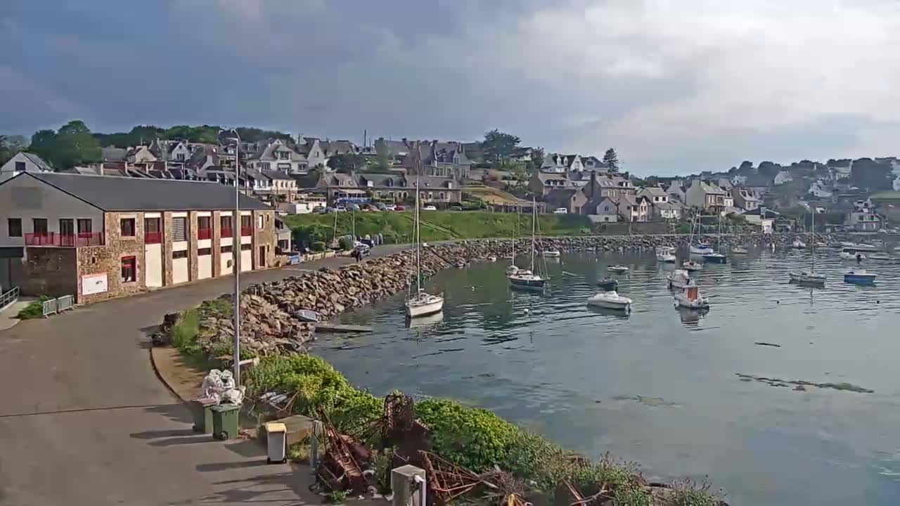 A calm harbor filled with sailboats is nestled next to a seaside town under a partly cloudy sky.