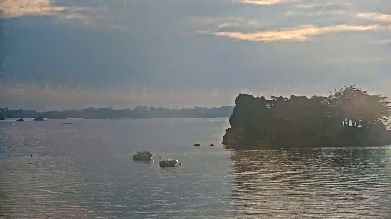 Two small boats are moored in calm water near a rocky islet topped with trees under a partly cloudy sky.