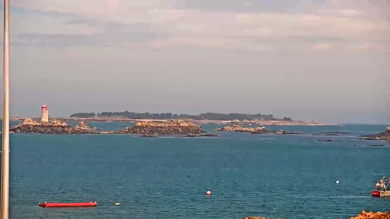 A red and white lighthouse stands on a rocky island in a calm, blue ocean under a mostly cloudy sky.