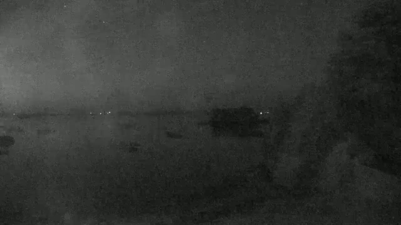 A nighttime view shows a dark body of water with indistinct shapes and a few faint lights in the distance under a cloudy sky.
