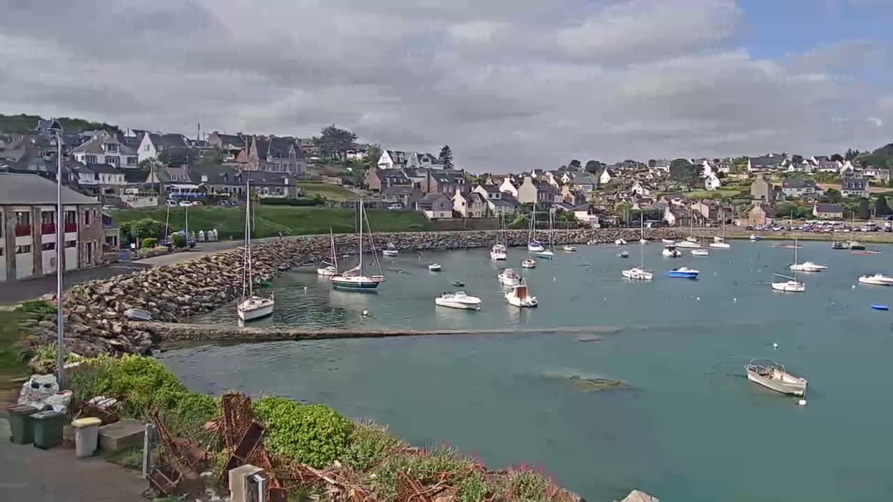 A small, calm harbor filled with sailboats and motorboats is nestled beside a coastal town under a partly cloudy sky.