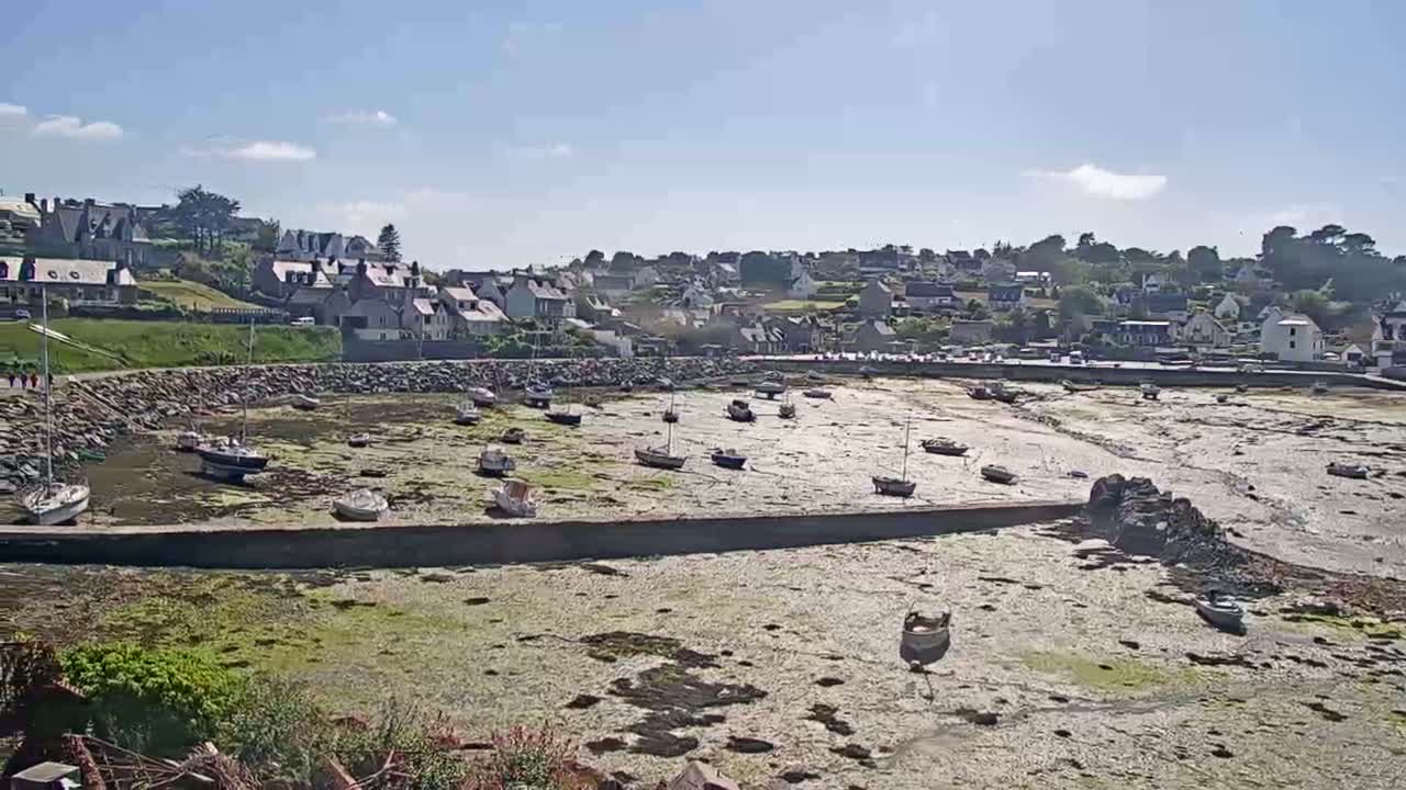 Numerous small boats sit on the exposed seabed of a harbor at low tide, in front of a small coastal town on a sunny day.