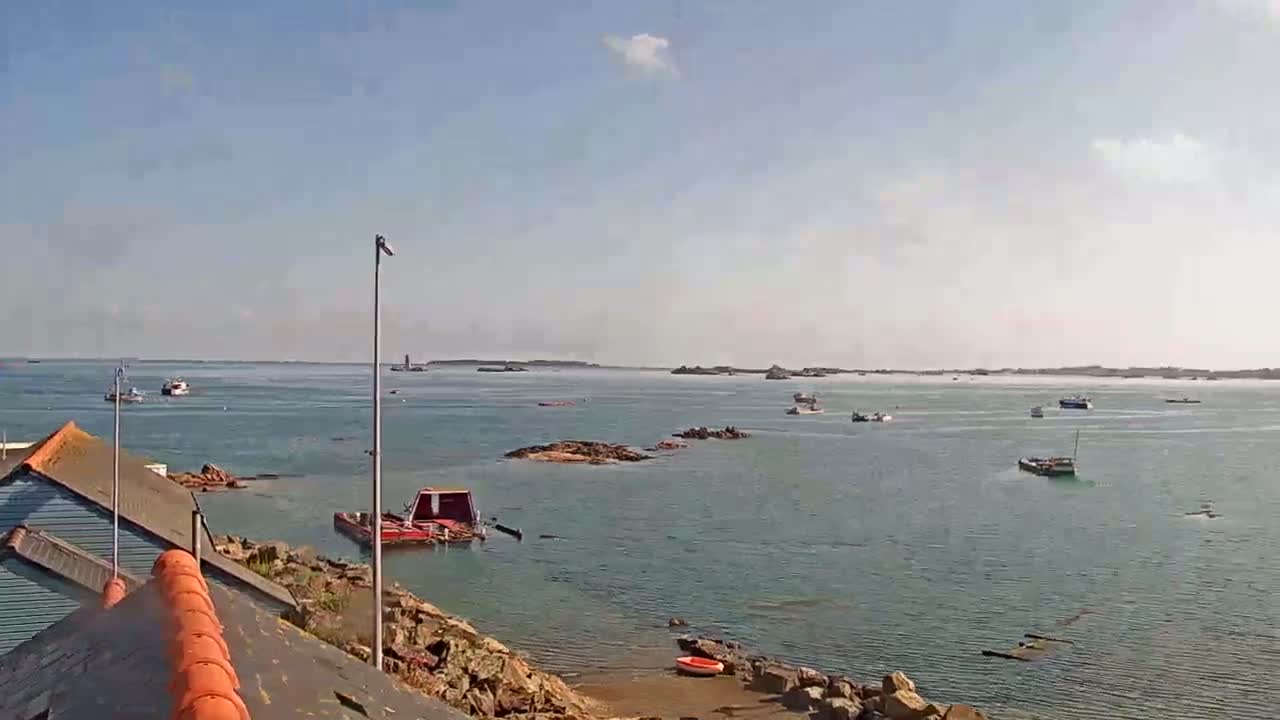 A calm, sunny day reveals a coastal scene with numerous small boats scattered across the water, near rocky outcrops and a small harbor, with a building partially visible on the left.