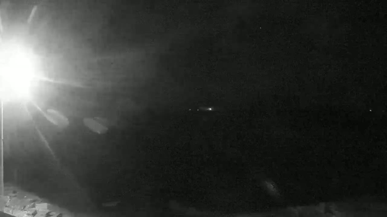 A bright light source shines in the lower left corner of a dark, nighttime scene, illuminating some ground in the foreground and casting light across a hazy, dark background with a few distant lights.