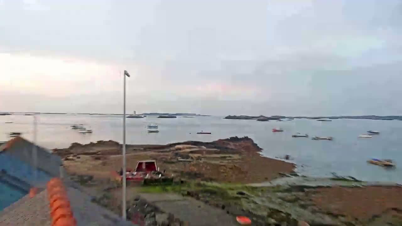 A calm, overcast day reveals a rocky shoreline with several small boats scattered across a bay, beyond which lie more distant islands.