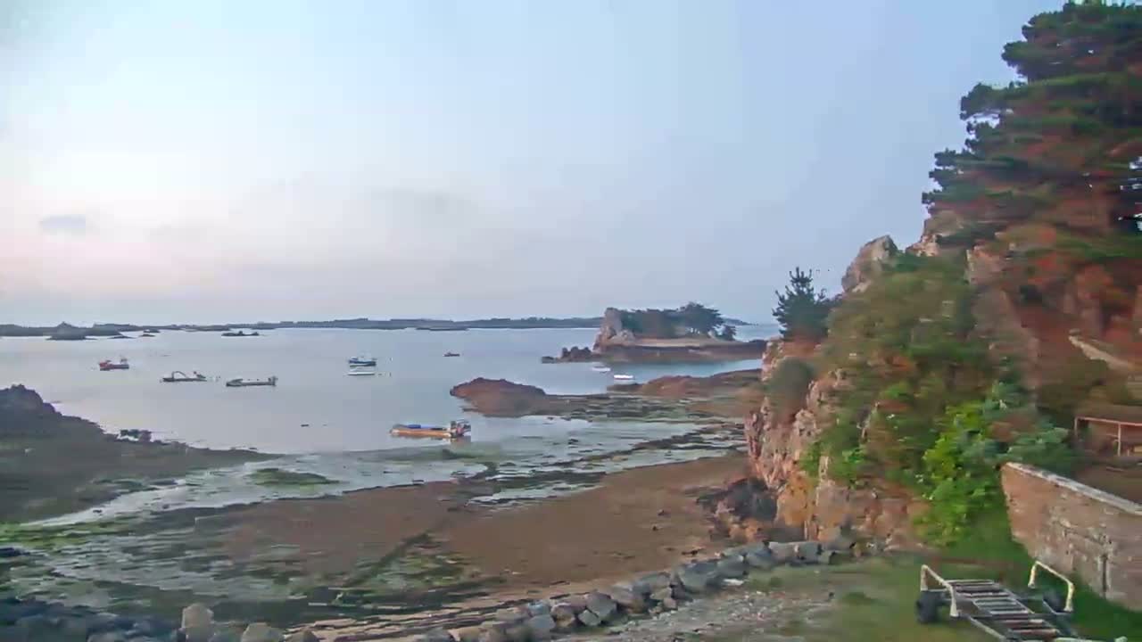 A calm, pale-blue bay dotted with small boats is partially enclosed by rocky, vegetated shores under a clear, light sky.