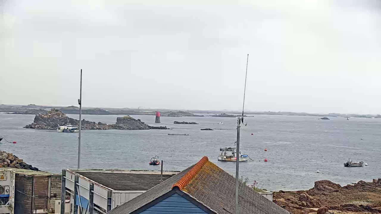 Several fishing boats are in a calm, gray-weather harbor near rocky islands and a red and white lighthouse.
