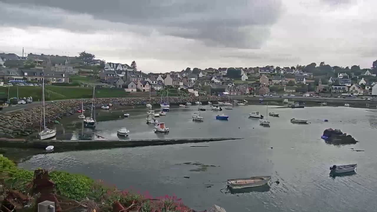 A small harbor filled with sailboats and motorboats is nestled below a hilltop village under an overcast sky.