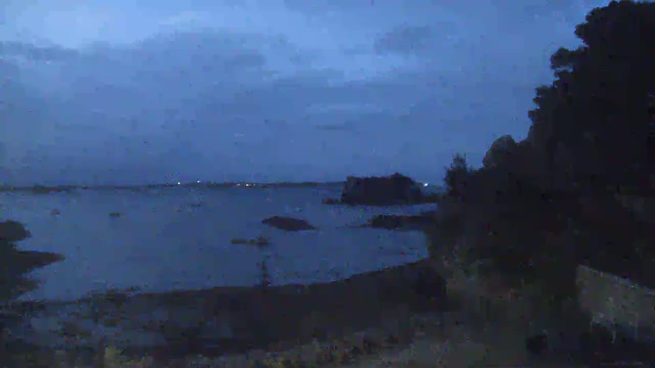 A dark, calm body of water is visible at twilight, with a shoreline, some small islands, and dark trees in the foreground.