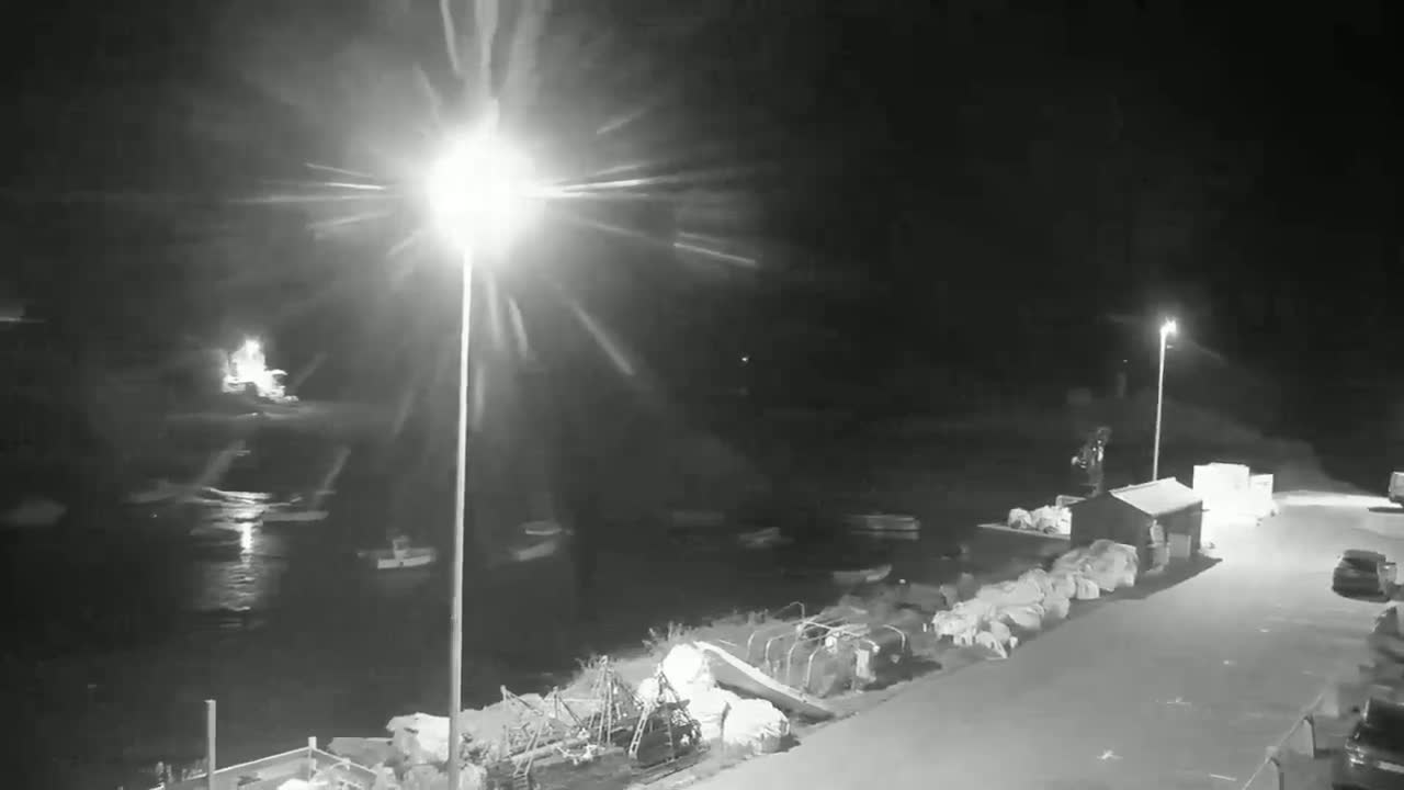 A nighttime, low-light view of a harbor with several boats moored, a small building, and a roadway, illuminated by bright lights.