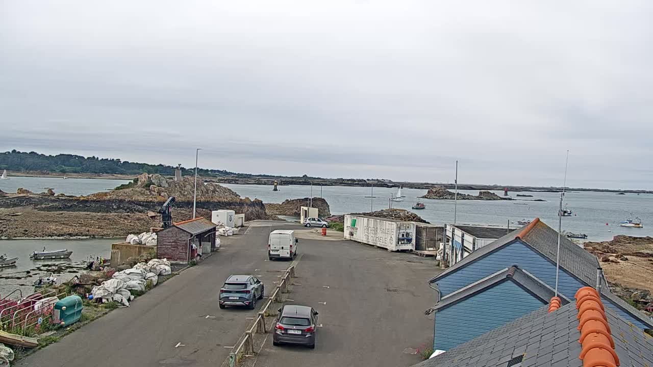 A harbor with several cars parked, small buildings, and boats in the water under an overcast sky.