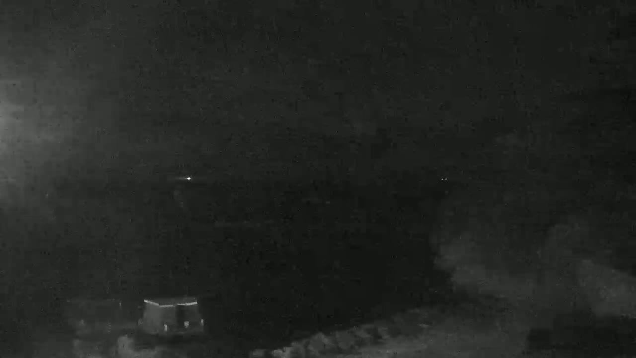 A nighttime, low-light image shows a dark, mostly indistinct landscape with a few faint lights visible in the distance.