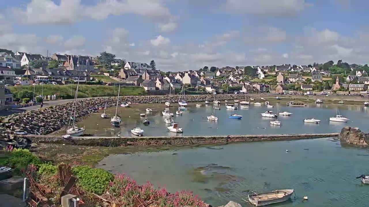 A small harbor filled with sailboats and motorboats is nestled beside a hillside town under a partly cloudy sky.