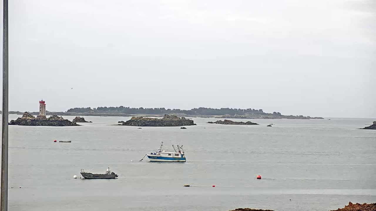 A calm, overcast day reveals a coastal scene with a lighthouse on a rocky islet, several small boats on the water, and a larger island in the distance.