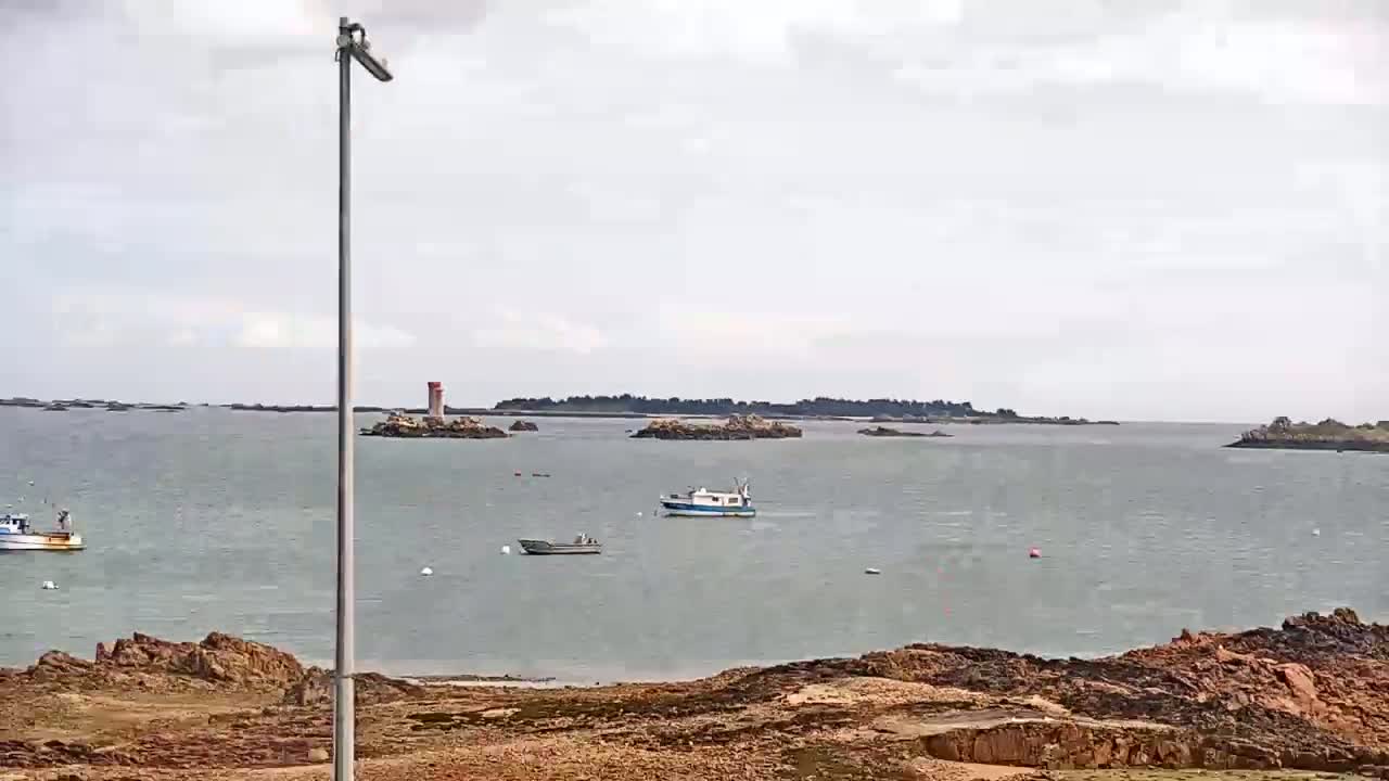 A calm, overcast day shows a body of water with several small boats and a lighthouse in the distance, near a rocky shoreline.