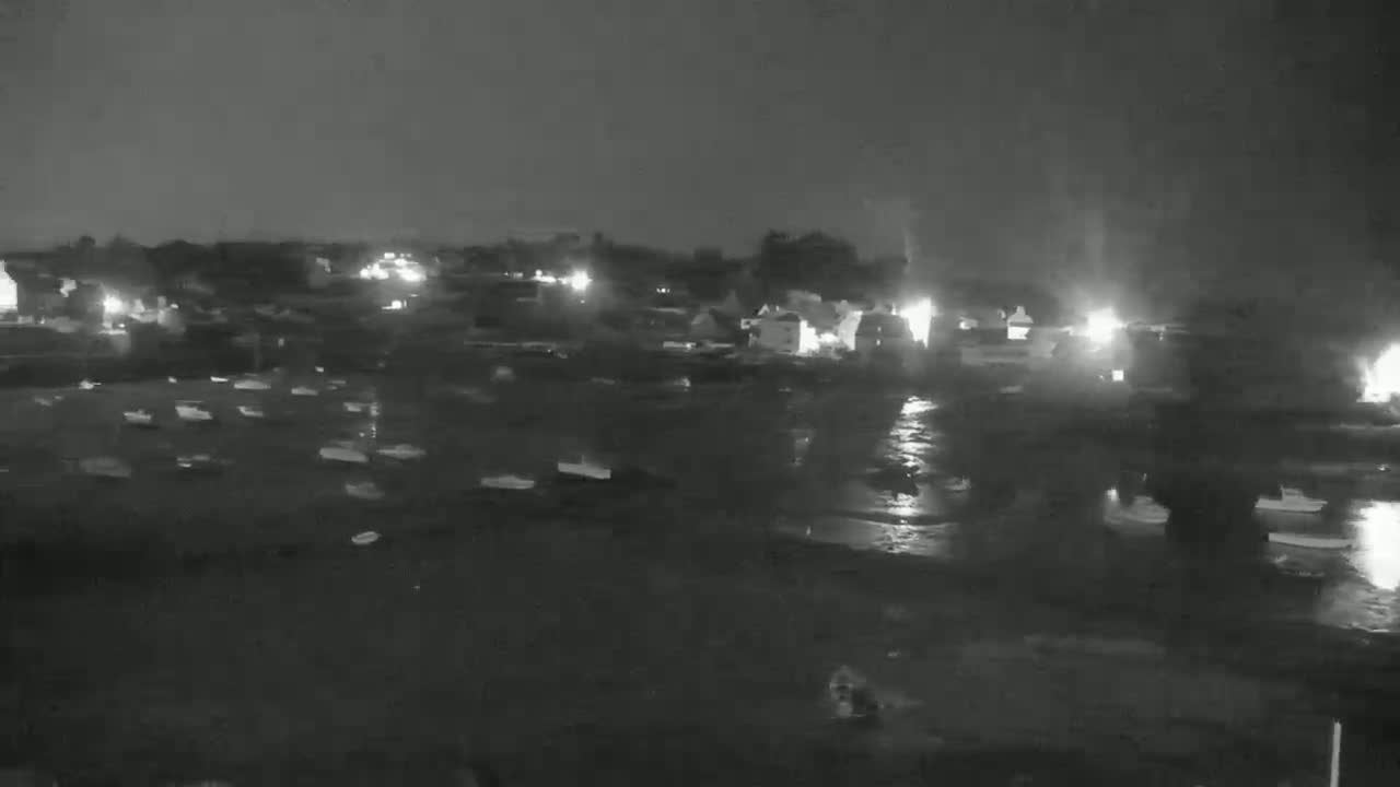 A nighttime, low-light view of numerous boats moored in a harbor, with a small town or village visible in the background under a dark, possibly overcast sky.