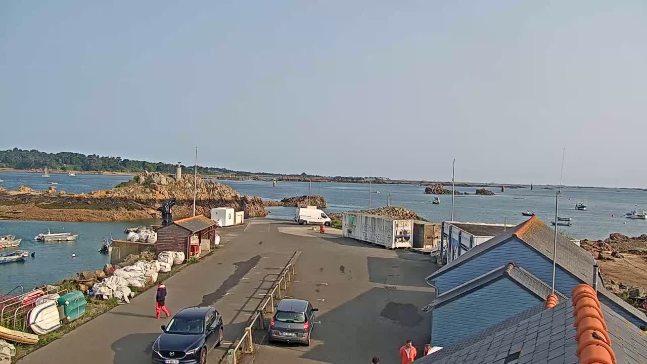 A sunny harbor scene shows several small boats moored near rocky islands, with cars parked in a lot near some buildings and cargo containers.