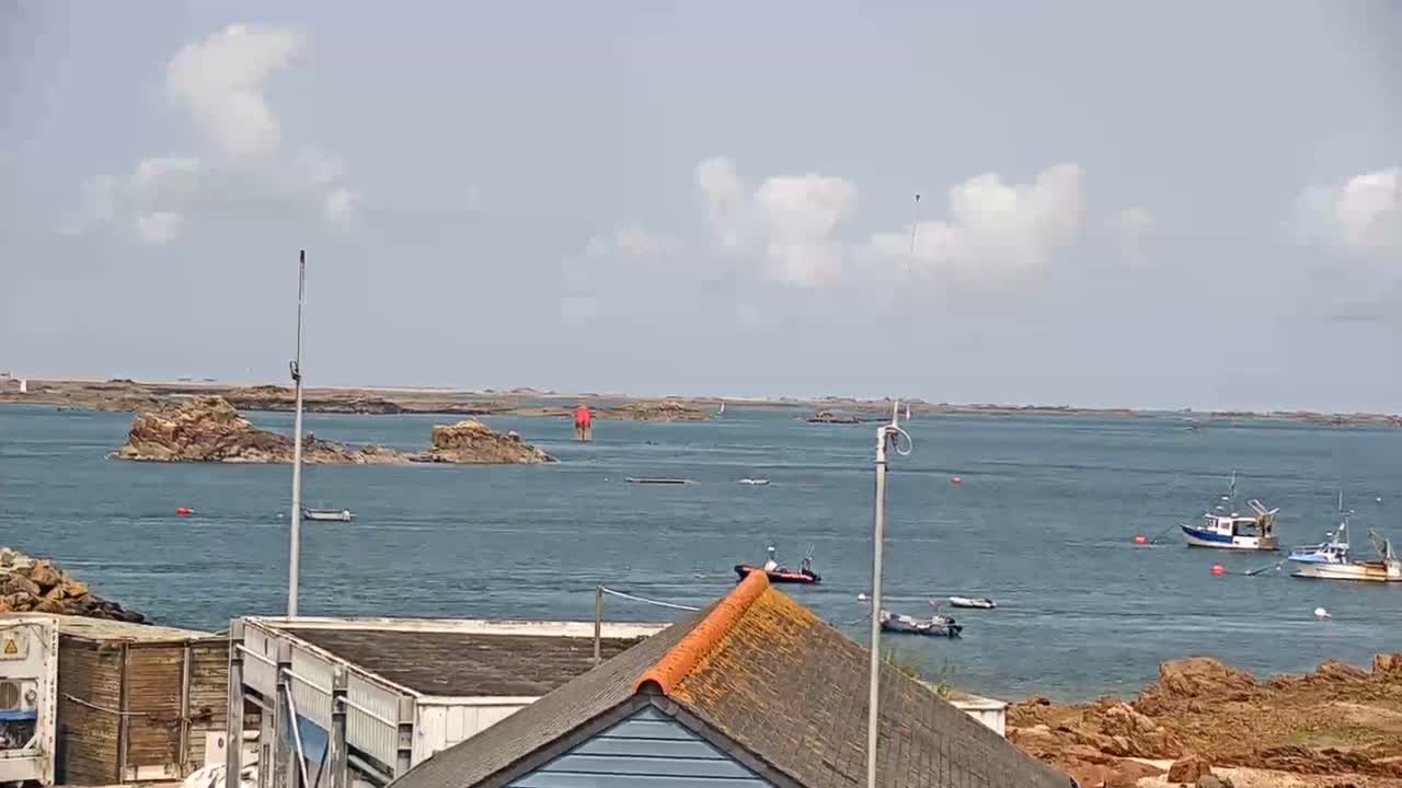 A partly cloudy day shows a calm body of water with several small boats and rocky islands in the distance, viewed from behind a building with an orange-tiled roof.