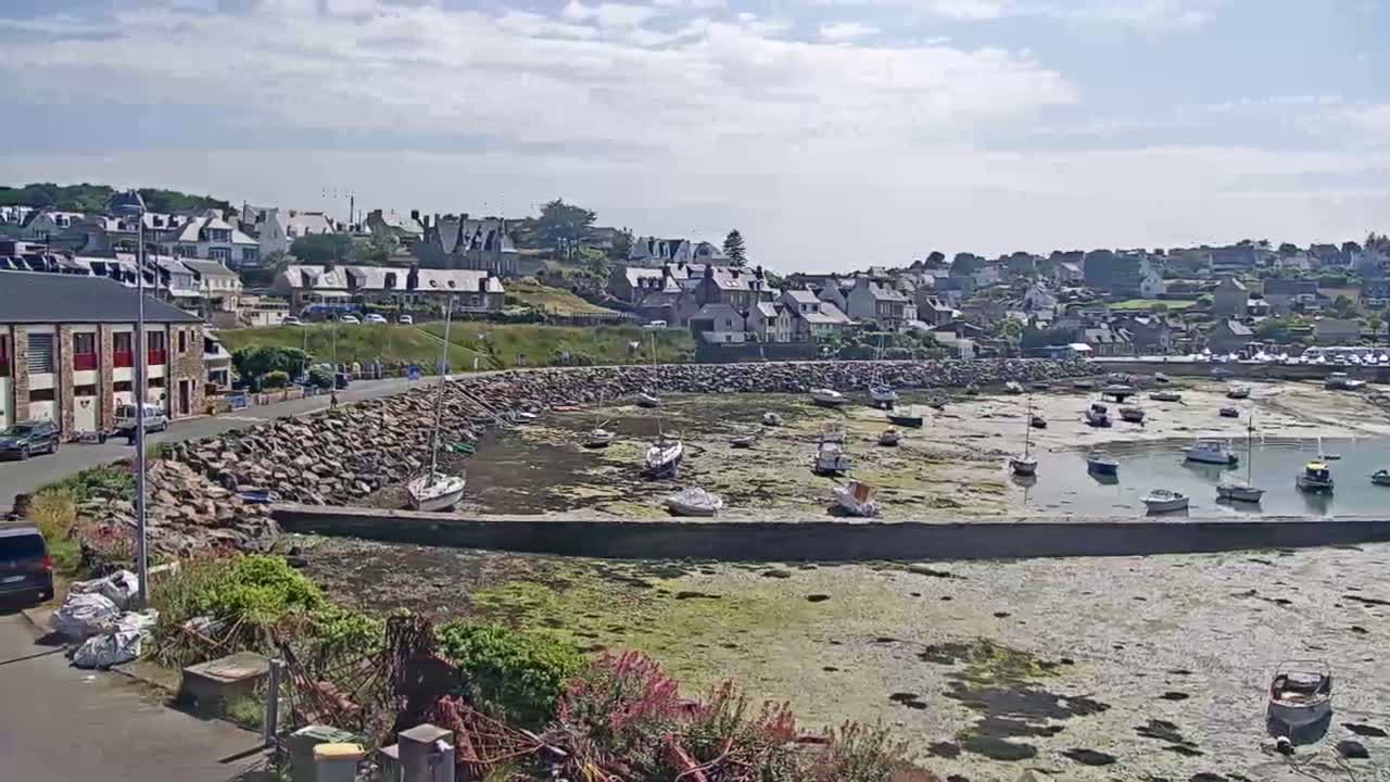 A small harbor with numerous boats sits partially exposed at low tide in front of a town under a partly cloudy sky.