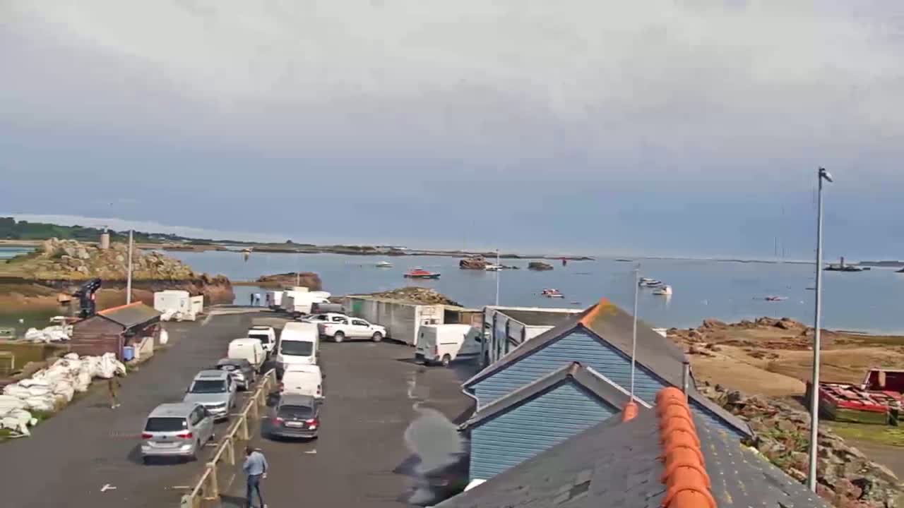 A harbor with several cars and vans parked near light blue buildings, overlooks calm water with a few boats, under a mostly cloudy sky.