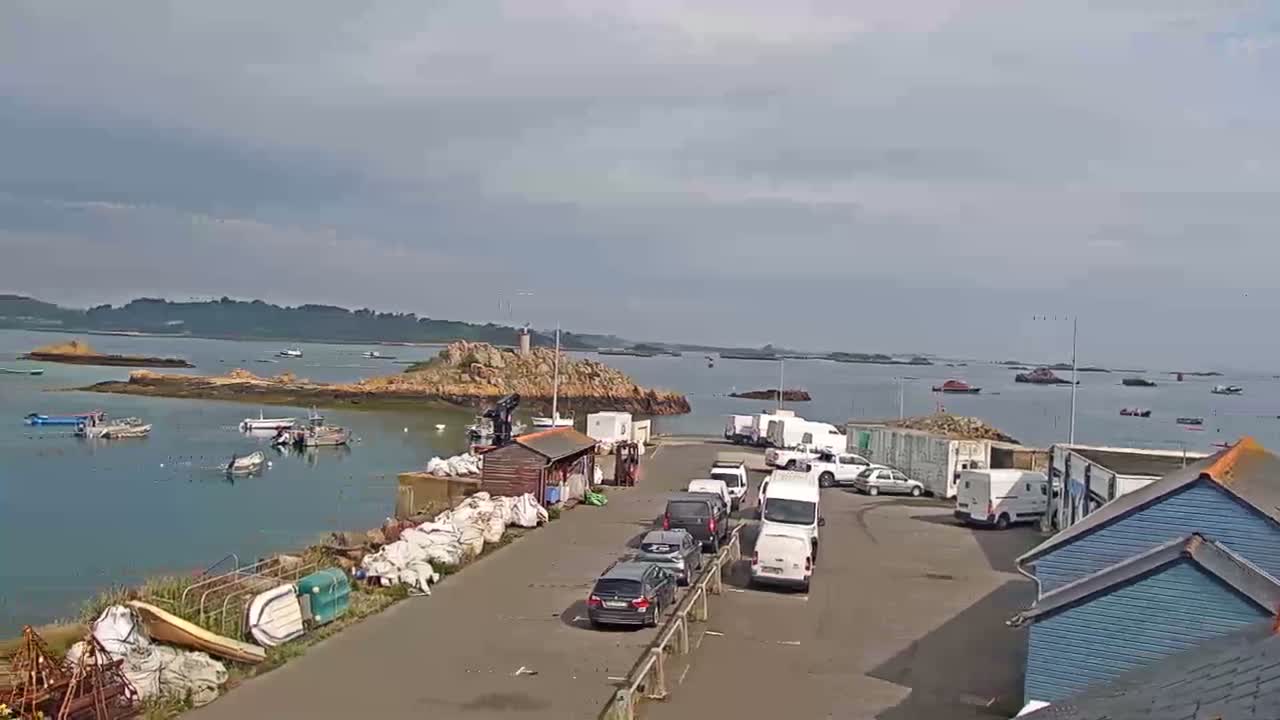 A harbor with several boats and cars parked near some small buildings, under a mostly cloudy sky.