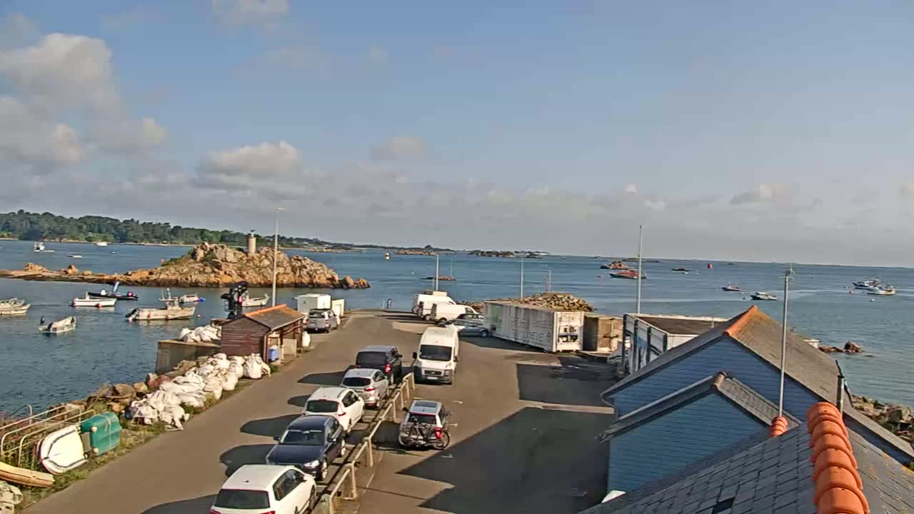A sunny day at a harbor shows several small boats at anchor near a rocky island, with cars parked in a lot next to several buildings.