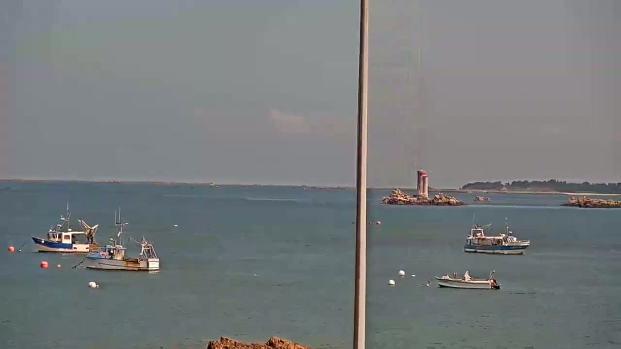 Several small fishing boats are anchored in calm, gray water near a small, rocky island with a lighthouse on a mostly clear day.