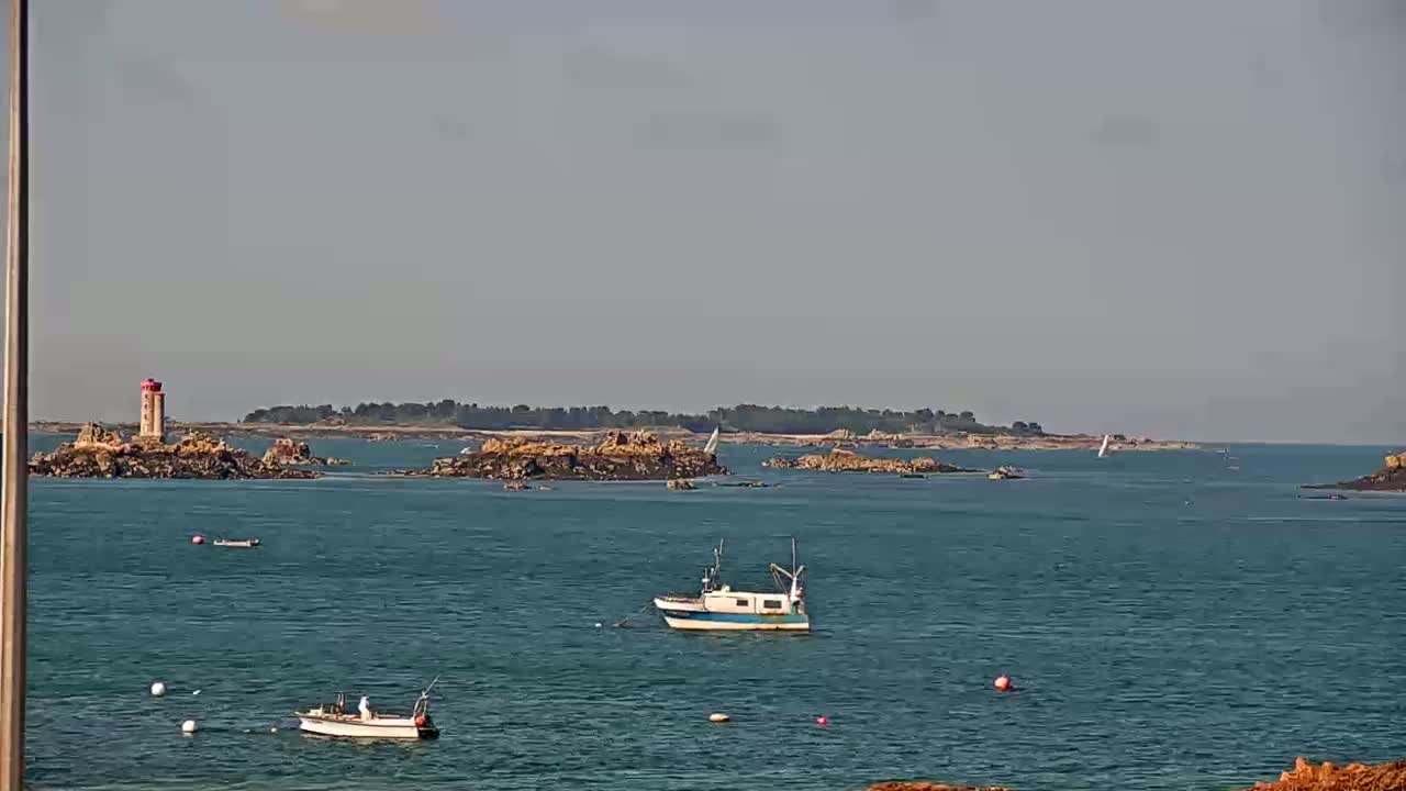A calm, hazy day shows a rocky island with a lighthouse, several small boats on the water, and a distant shoreline.