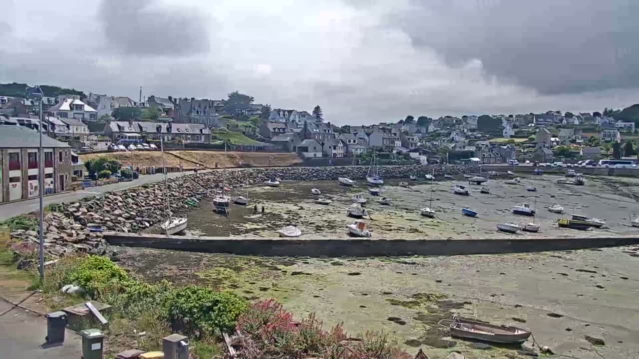 A small harbor filled with numerous boats sits at low tide under a mostly cloudy sky, with a village of houses visible on a hillside in the background.
