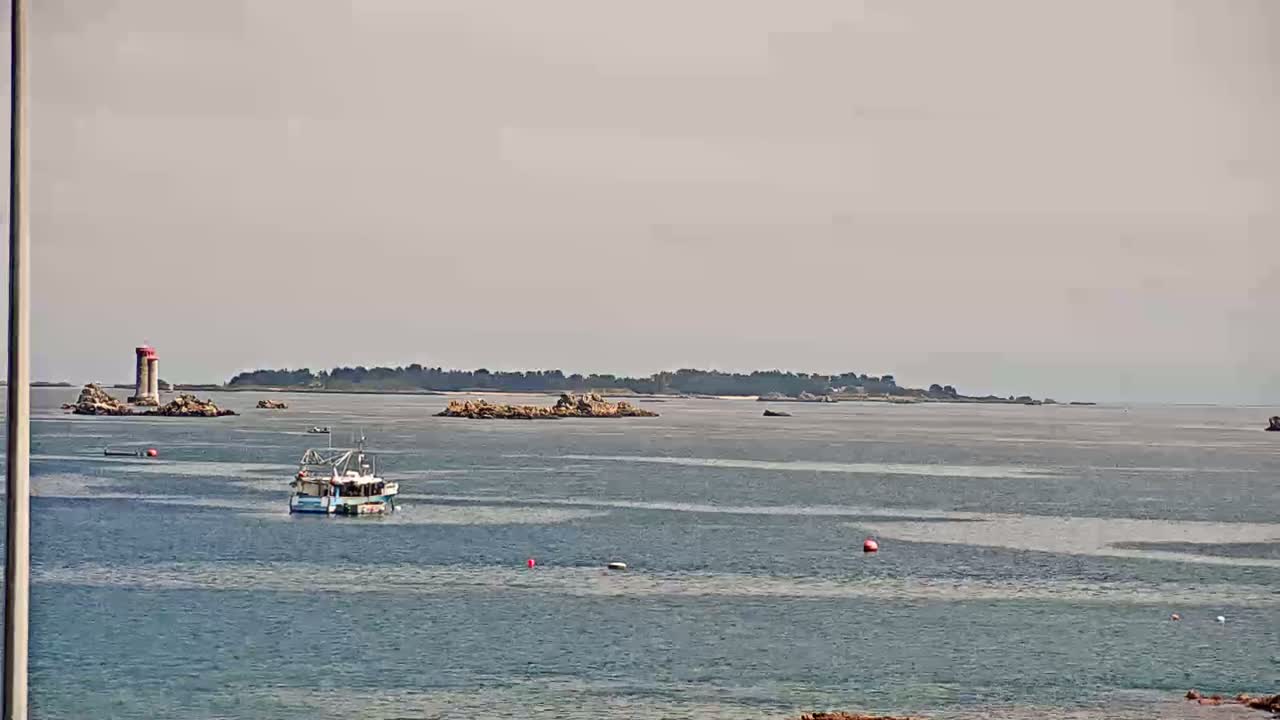 A small fishing boat is on calm, light-blue water near rocky islands and a lighthouse under an overcast sky.